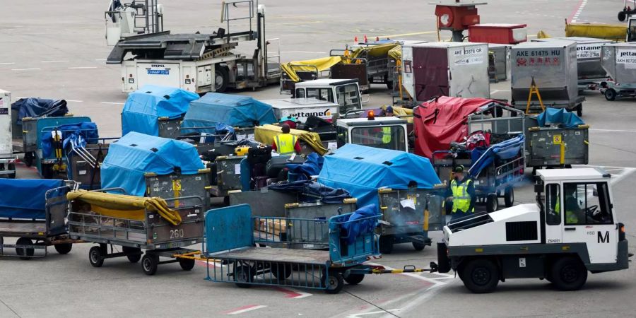 Ein leerer Gepäcktransportwagen fährt auf einem Vorfeld an zahlreichen mit Gepäck beladenen Wagen vorbei.