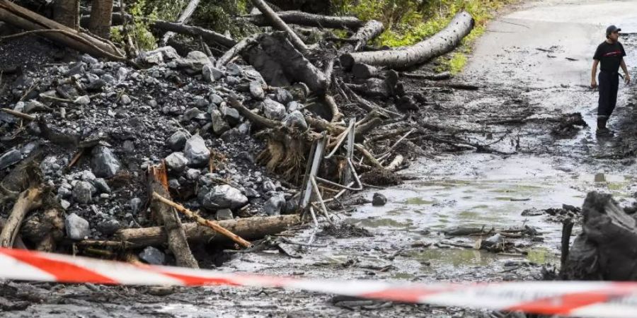 Ein Mann steht auf einer abgesperrten Strasse, die von Schlamm, Bäumen und Geröll bedeckt ist. Nach einem heftigen Regen wurde ein Auto durch das Hochwasser des Baches Losentze mitgerissen. In dem Auto befanden sich zwei Menschen, die noch als vermisst gelten. Foto: Jean-Christophe Bott/KEYSTONE