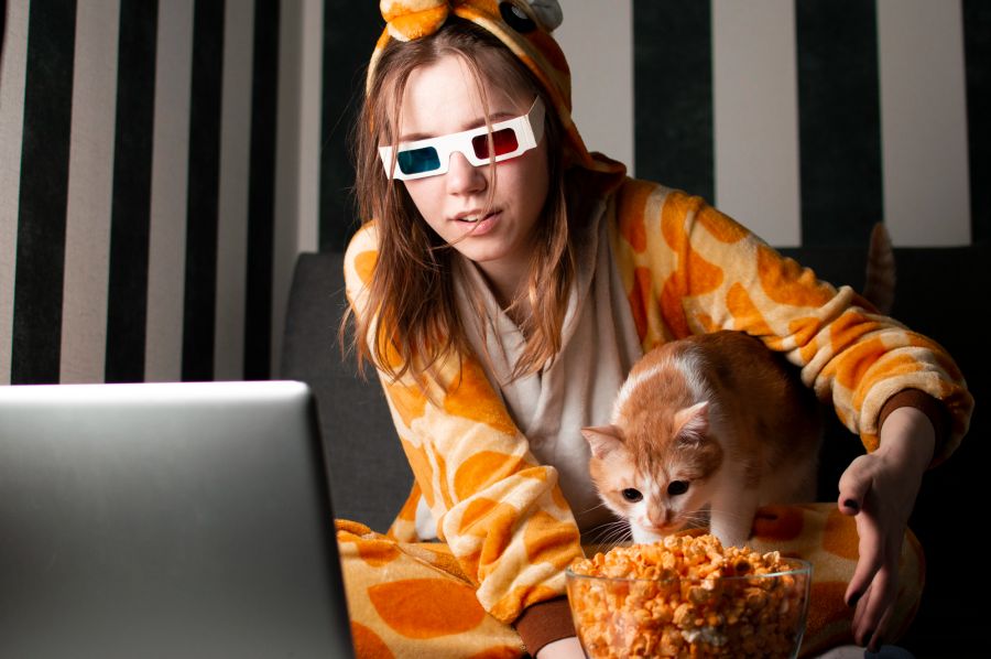 Junge Frau mit Kinobrille und ihre Katze, die aus einer Popcornschüssel fressen will