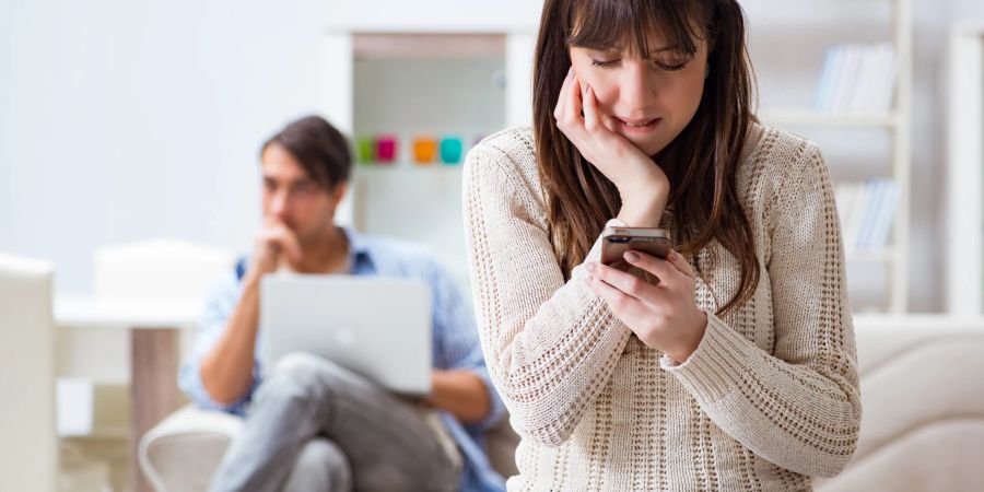 frau schaut mit zusammen gekniffenen zähnen auf ihr handy, mann mit laptop im hintergrund