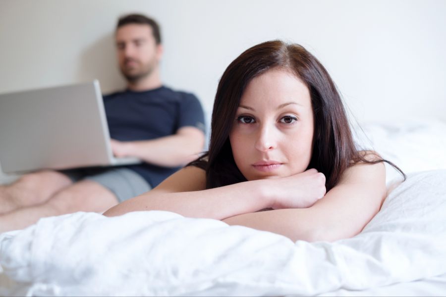 Nachdenkliche Frau liegt auf einem Bett, Mann im Hintergrund arbeitet am Laptop