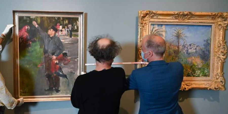 Martin Faass (r), Direktor des Hessischen Landesmuseums Darmstadt, die Restauratorin Olivia Levental (l) und Depotverwalter Wolfgang Koch bereiten Liebermanns "Der Papageienmann (1900/01)"neben dem Gemälde "Villen in Bordighera (1884)" von Claude Monet für die Hängung vor. Foto: Arne Dedert/dpa