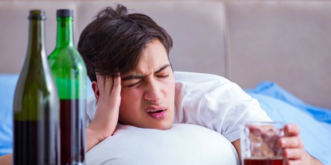 Mann im Bett hält sich den Kopf, daneben Weinflaschen und ein Glas mit Alkohol.