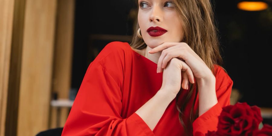 Junge Frau mit roten Lippen und rotem Kleid schaut in die Ferne, vor ihr rote Rosen.