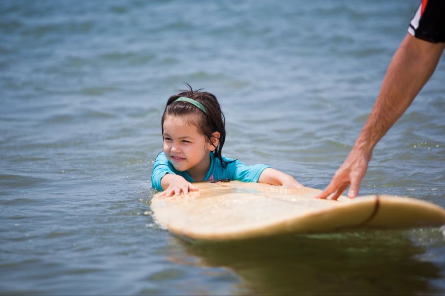 Kleines Mädchen im Meer hält sich an Surfboard fest.