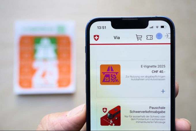 Ein Mann hält ein Smartphone mit der E-Vignette für das Jahr 2025, im Hintergrund eine herkömmliche Autobahnvignette. E-Vignette