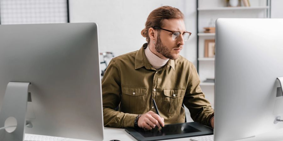 Mann sitzt am Schreibtisch zwischen zwei Monitoren