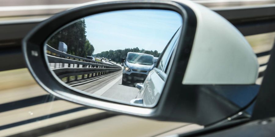 Im Aussenspiegel eines Pkw, der etwa 120 Kilometer pro Stunde und auf der linken Fahrbahn fährt, ist ein Kleintransporter zu sehen, der viel zu dicht auffährt und keinen Sicherheitsabstand einhält.