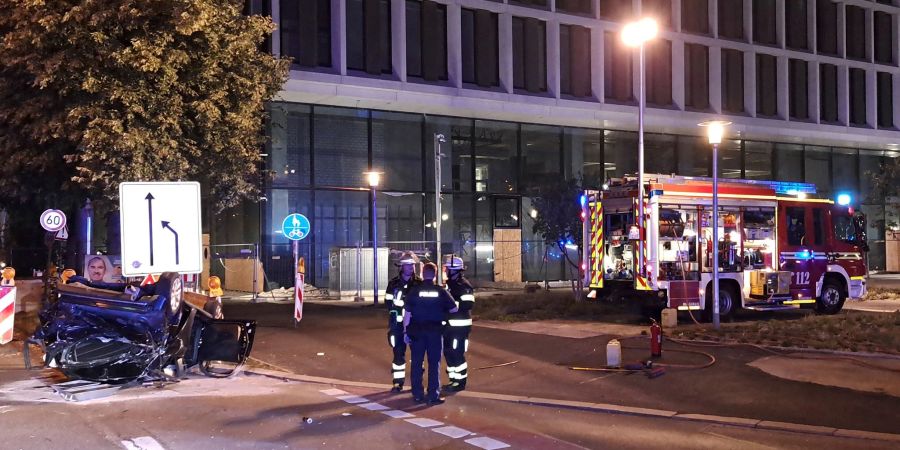 Einsatzkräfte der Feuerwehr und Polizei stehen neben einem zerstörtem Wagen an der Unfallstelle in München.