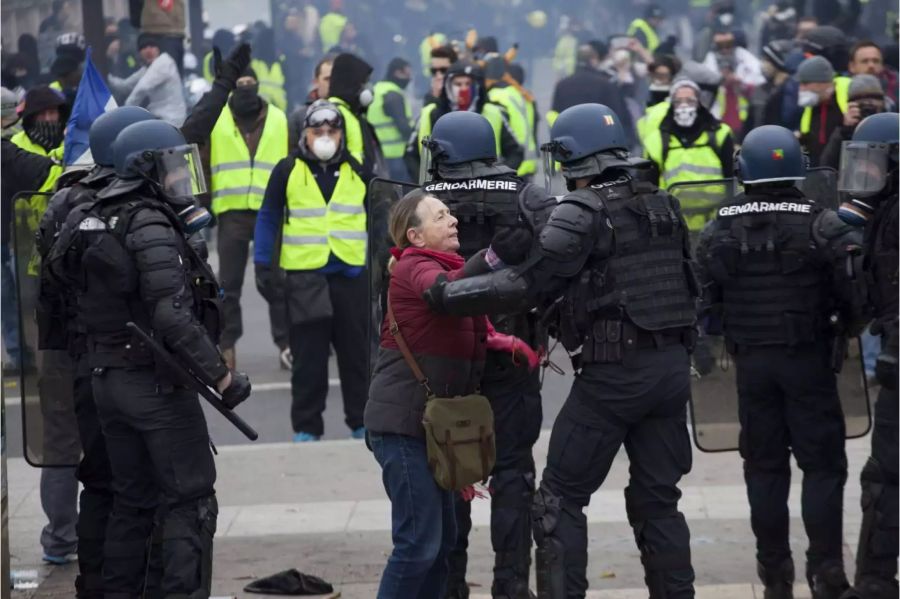 Vermummte Demonstranten stehen Polizisten gegenüber, von denen einer mit einer älteren Frau spricht.