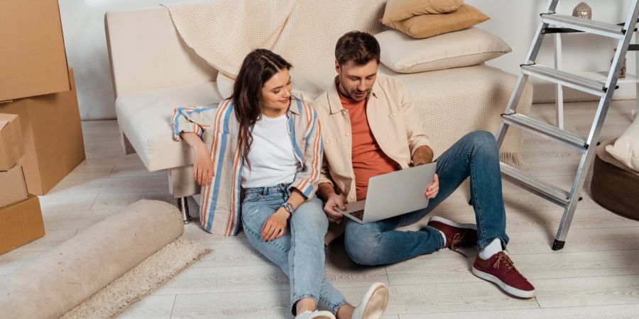 Mann und Frau sitzen im Wohnzimmer mit einem Laptop an ein Sofa angelehnt