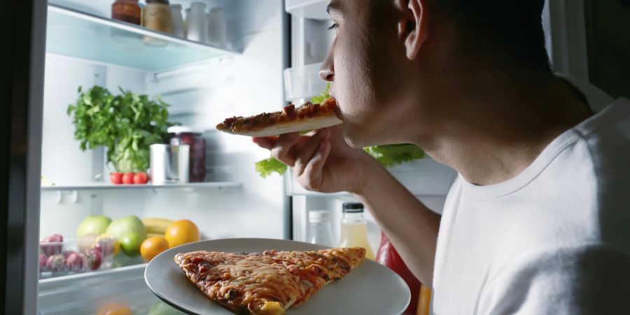 Mann isst Pizza vor dem Kühlschrank