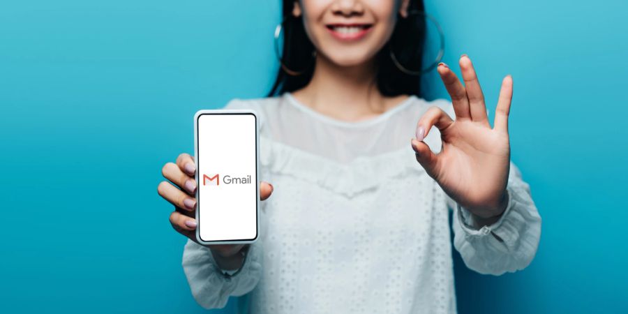 Eine Frau hält ein Smartphone in der linken Hand, auf dem das Logo von Gmail angezeigt wird. Mit der rechten Hand zeigt sie eine Okay-Geste.