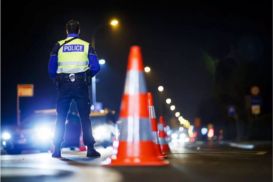 Polizist steht neben Strassenhüttchen vor einer zu kontrollierenden Schlange von Autos - Keystone