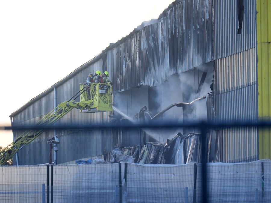 Feuerwehrleute auf einer Hebebühne kühlen die ausgebrannte Lagerhalle ab – das Ausmass der Zerstörung ist deutlich sichtbar.
