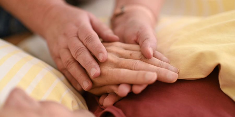 Eine Hospizmitarbeiterin hält die Hand eines todkranken Menschen, der in einem Hospiz im Bett liegt.