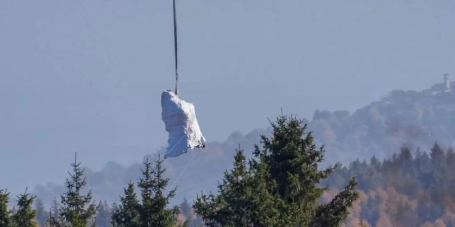 Ein Hubschrauber der italienischen Feuerwehr fliegt die in weisse Planen gehüllte Kabine der abgestürzten Mottarone-Seilbahngondel vom Lago Maggiore ins Tal. Foto: Antonio Calanni/AP/dpa