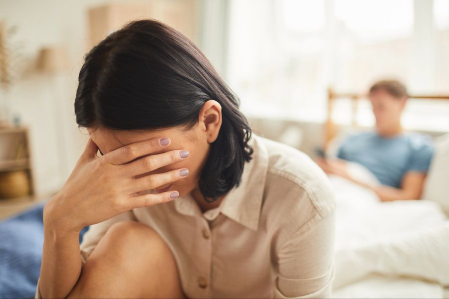 Verzweifelte Frau hält sich Hand vor das Gesicht, Mann im Hintergrund im Bett am Smartphone