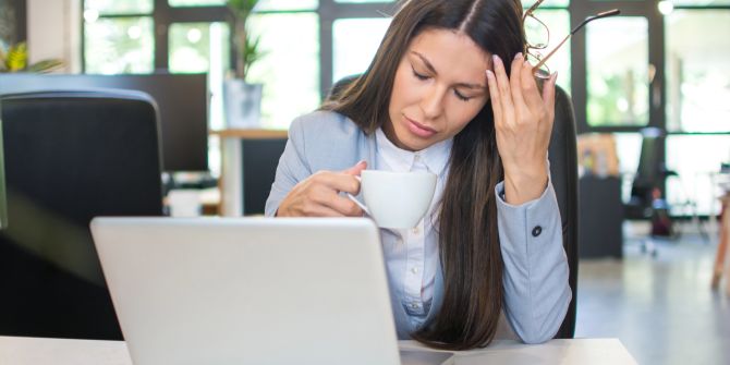 Erschöpfte Frau im Büro mit Kaffeetasse am Laptop