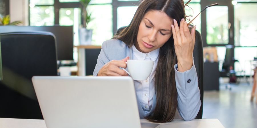 Erschöpfte Frau im Büro mit Kaffeetasse am Laptop