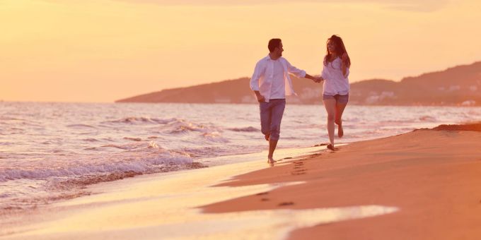 Für eine romantische Reise sind sowohl Städtetrips als auch Ausflüge in die Natur und ans Wasser geeignet. Pärchen läuft glücklich am Strand entlang
