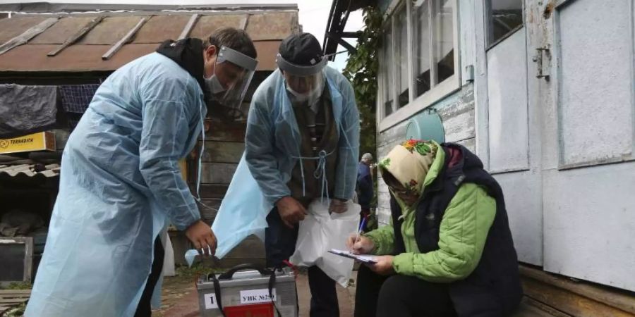 Zwei Wahlhelfer stehen neben einer Frau, die auf den Stufen vor einem Haus sitzt und Dokumente ausfüllt. Foto: Evgeniy Sofiychuk/AP/dpa