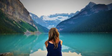 Frau sitzt an einem sehr blauen See und schaut in die Berge in der Ferne.