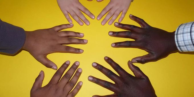 Drei Personen unterschiedlicher Hautfarbe halten ihre Hände zu einem Kreis auf einem gelben Tisch. Integration