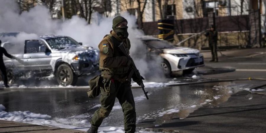 Ein ukrainischer Soldat bezieht Stellung vor einer Militäreinrichtung, während Feuerwehrleute im Hintergrund zwei brennende Autos löschen. Foto: Emilio Morenatti/AP/dpa