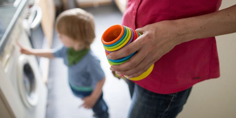 Eine Mutter hält Kinderspielzeug in der Hand, während ein kleiner Junge an einer Waschmaschine spielt. Die Krisen der letzten Jahre belasten vor allem Mütter - Stress und finanzielle Probleme sorgen für eine Dauerbelastung.