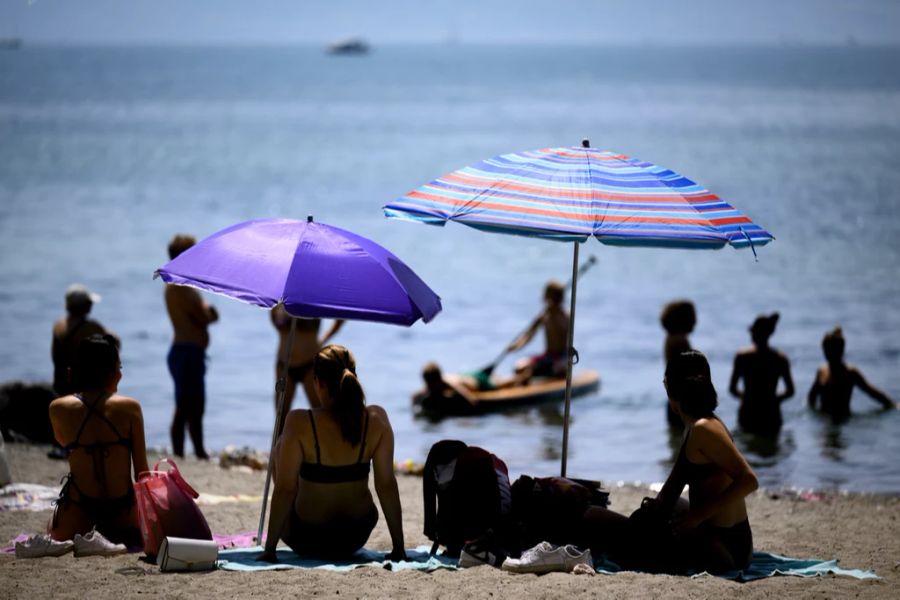 Menschen kühlen sich bei warmem Wetter im Schatten von Sonnenschirmen am Ufer des Genfer Sees am Strand von Vidy (Plage de Vidy) in Lausanne, Schweiz, am Sonntag, 19. Juni 2022.