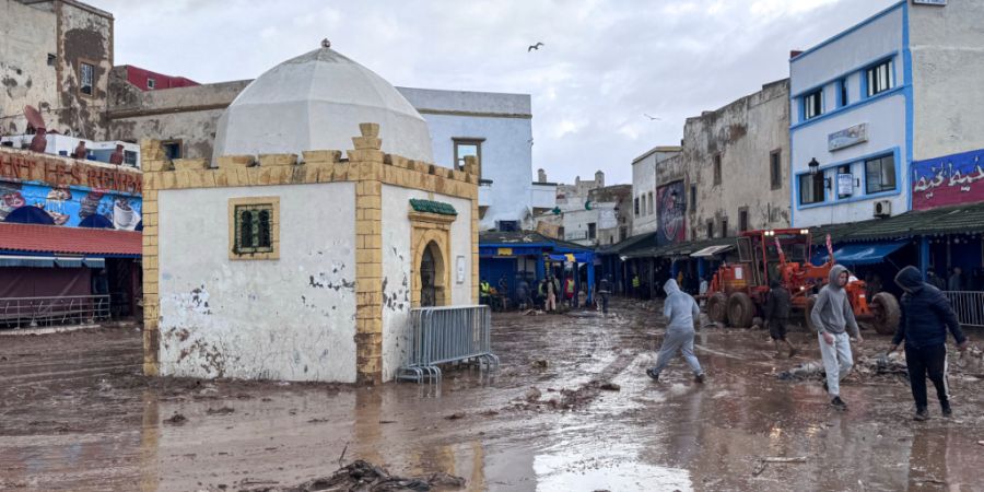 Menschen gehen auf einer von Matsch bedeckten Straße entlang. In Marokko sind nach schweren Regenfällen und Überschwemmungen mindestens 37 Menschen ums Leben gekommen. Foto: Abderraz...