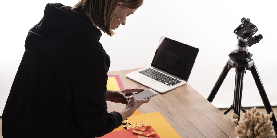 junge frau schaut auf smartphone, schreibtisch, kamera mit stativ und laptop im hintergrund