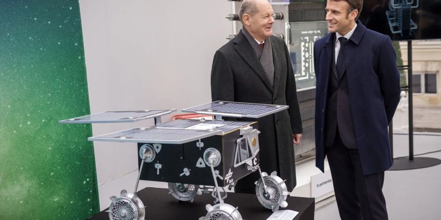 Bundeskanzler Olaf Scholz (SPD, l), und Emmanuel Macron, Präsident von Frankreich, stehen bei einer Industrieausstellung im Rahmen des Deutsch-Französischen Ministerrats hinter einem Rover, der gemeinsam von DLR und CNES gebaut wurde.