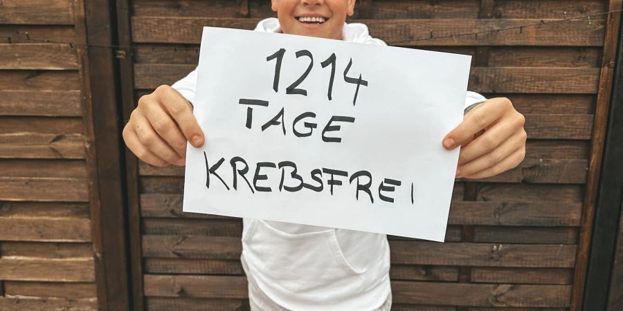 Der junge Lufthansa-Flugbegleiter zeigt im Dezember 2022 in einem Garten bei Köln ein Schild mit der Aufschrift «1214 Tage krebsfrei».