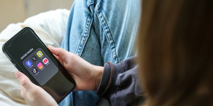 ILLUSTRATION - Ein Mädchen hält ein iPhone 16 in den Händen, auf dessen Display in einem Ordner mit der Bezeichnung «social media» die Logos von Facebook, X (ehemals Twitter), Snapch...