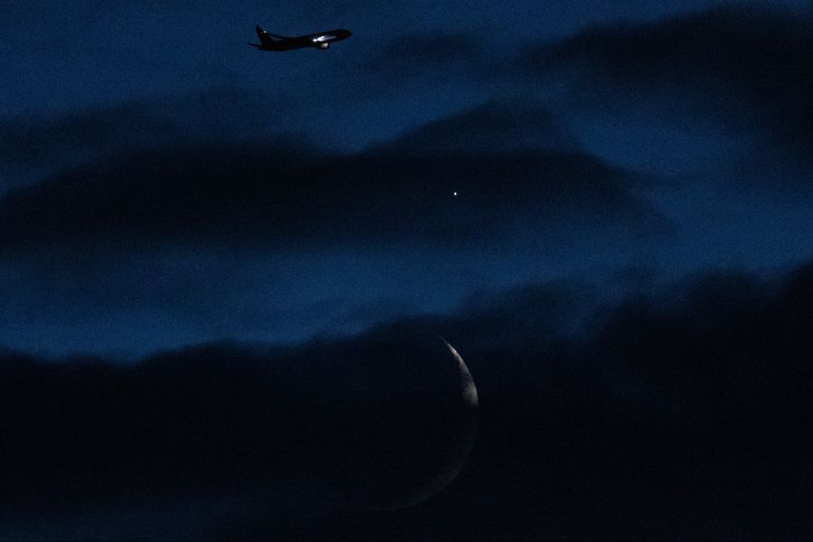 Ein Flugzeug fliegt am sichelförmigen Mond und der Venus hinter Wolken vorbei.