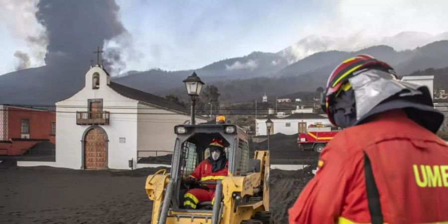 Einsatzkräfte des Militärs entfernen schwarze Asche vom Vulkan, der hinter der kleinen Kirche auf der Kanareninsel La Palma weiter Lava ausstösst. Foto: Saul Santos/AP/dpa