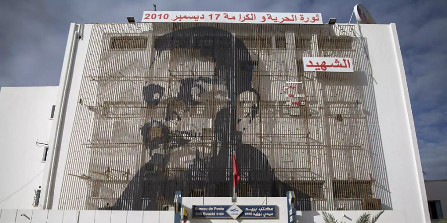 ARCHIV - Auf der Fassade eines Postamtes in der tunesischen Kleinstadt Sidi Bouzid ist das Gesicht des tunesischen Gemüsehändlers Mohammed Bouazizi abgebildet, der sich am 17. Dezember 2010 selbst anzündete - aus Verzweiflung über seine Lebenslage und die Willkür der Behörden. Foto: Riadh Dridi/AP/dpa