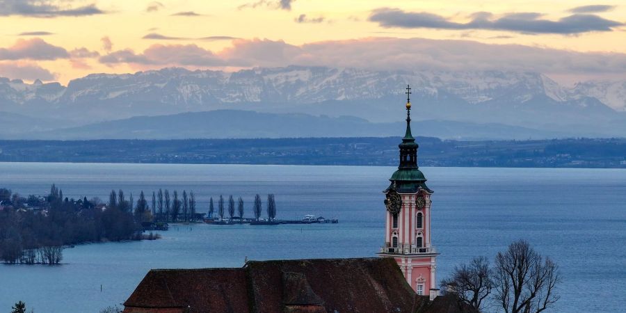 Hinter der ehemaligen Klosterkirche Birnau am Bodensee geht die Sonne auf. Die Alpen sind im Hintergrund zu sehen.