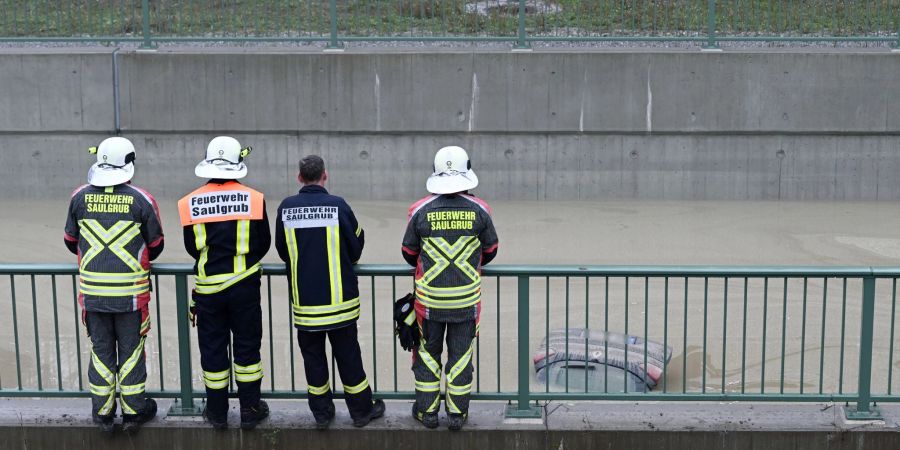 Einsatzkräfte der Feuerwehr schauen auf die Heckklappe eines Autos, das aus dem Wasser auf der teilweise überfluteten Bundesstrasse B23 ragt.
