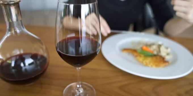 Ein Glas Rotwein steht auf einem Tisch, im Hintergrund isst eine Frau eine Mahlzeit von einem Teller. (Symboldbild) Coronavirus
