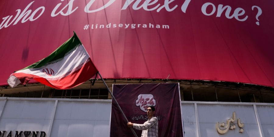 Ein Mann schwenkt eine iranische Flagge unter einer Werbetafel, auf der auf Englisch zu lesen ist: «Who is D nexT one?» und «#lindseygraham». Foto: Vahid Salemi/AP/dpa