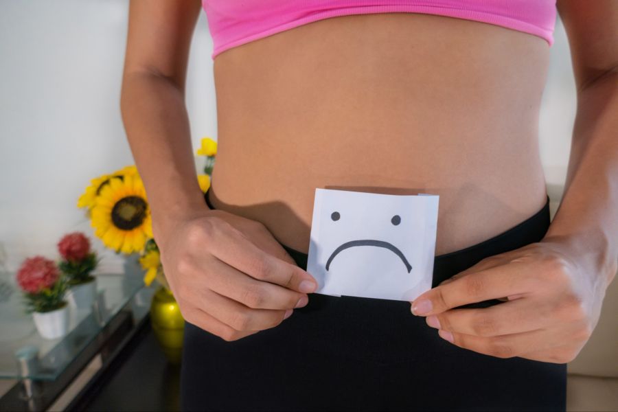 Frau hält traurigen Smiley in der Hand vor dem Intimbereich